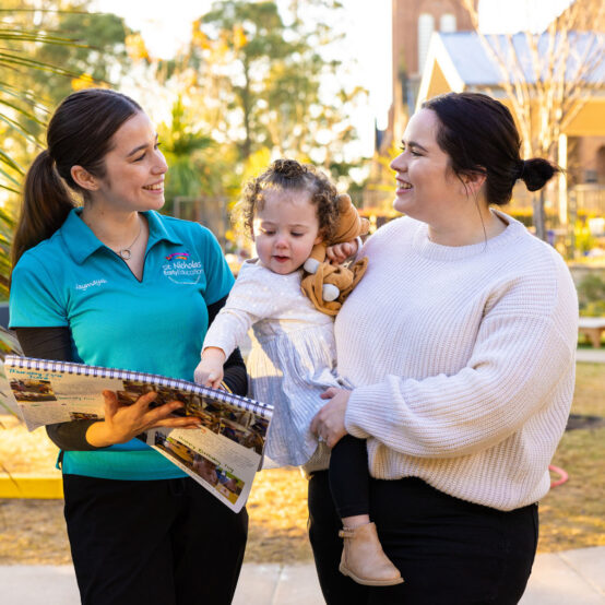 Child and parent on a centre tour with an educator in a teal shirt. Child is pointing to something in a floor book