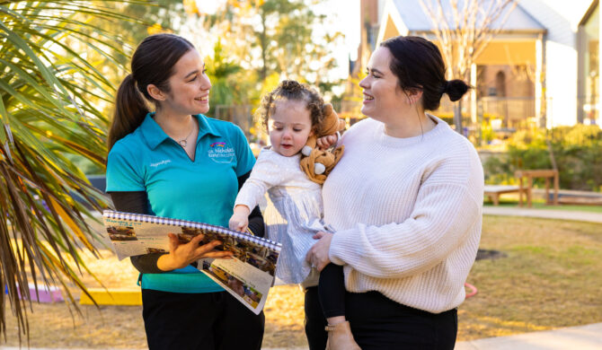 Child and parent on a centre tour with an educator in a teal shirt. Child is pointing to something in a floor book